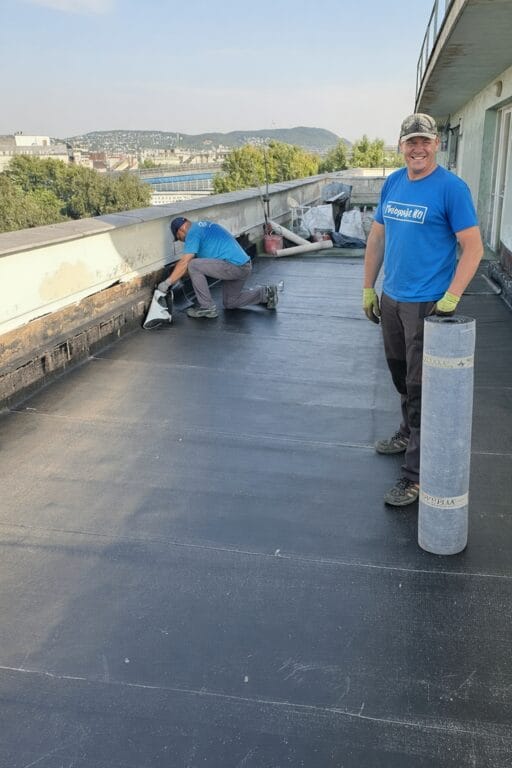 Két munkatárs sötét tetőlemezzel dolgozik egy lapostető szélén, mögöttük a város
