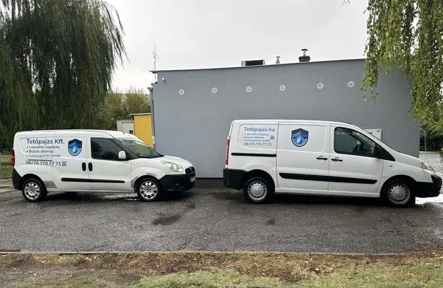 Két Tetőpajzs feliratú kisteherautó szürke épületfal mellett